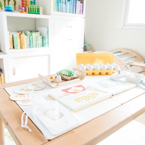 May include: A wooden table with a white tablecloth set for a pretend play breakfast. There are toy eggs, a toy frying pan, a toy spatula, a toy bread, a toy avocado, and a book that says "eggs" on it. There are also some cards with pictures of food on them.