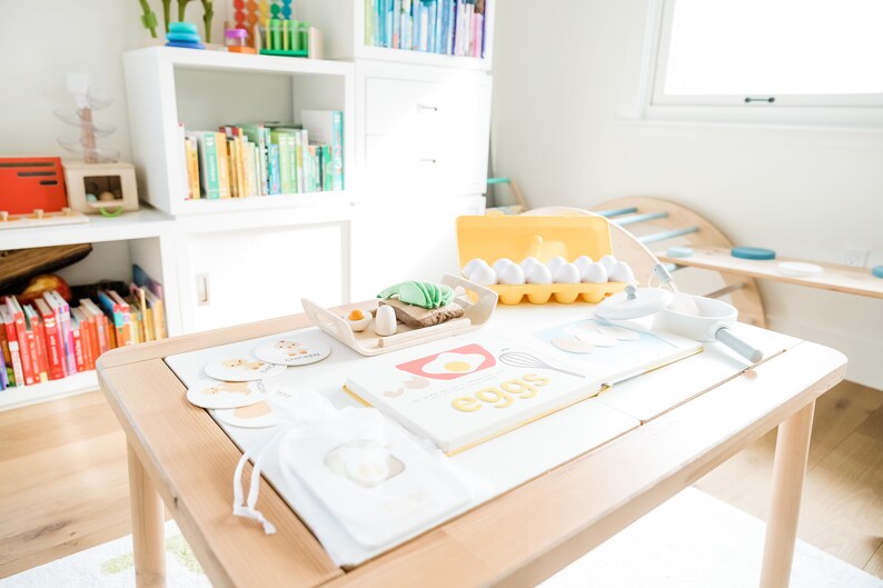 May include: A wooden table with a white tablecloth set for a pretend play breakfast. There are toy eggs, a toy frying pan, a toy spatula, a toy bread, a toy avocado, and a book that says "eggs" on it. There are also some cards with pictures of food on them.