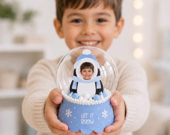 Könnte beinhalten: Eine transparente, kugelförmige Schneekugel mit einer Pinguinfigur, die das Foto eines Kindergesichts zeigt. Der hellblaue Sockel ist mit weißen Schneeflocken und dem Schriftzug "LET IT SNOW" verziert. Im Inneren befinden sich weiße Schaumstoffkugeln.