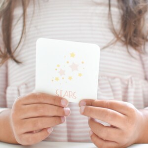 May include: A white card with pink and yellow stars and the word "STARS" printed on it. The card is being held by a child's hands.