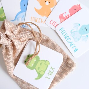 K&ouml;nnte beinhalten: Ein Dinosaurier-Memory-Spiel mit bunten Illustrationen von Dinosauriern, darunter ein T-Rex, ein Triceratops, ein Stegosaurus und ein Pterodactylus. Die Karten sind in einem kleinen Jutesack mit Kordel verpackt.