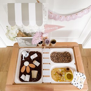 May include: A wooden sensory table with three white plastic bins filled with various items. The bins contain brown beans, small yellow beads, and a mix of items like a watering can, wooden flowers, and small cards. A banner that says "Showers" is above the table.