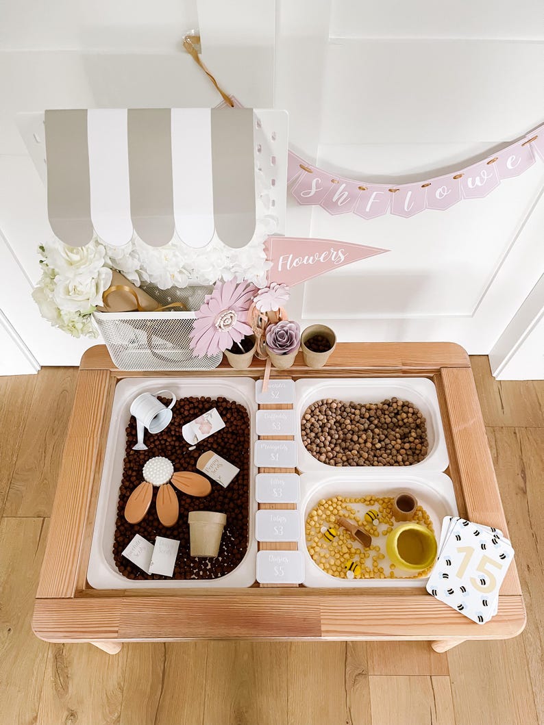 May include: A wooden sensory table with three white plastic bins filled with various items. The bins contain brown beans, small yellow beads, and a mix of items like a watering can, wooden flowers, and small cards. A banner that says "Showers" is above the table.