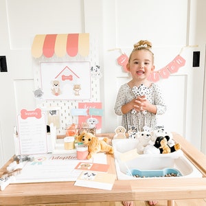 May include: A child plays with a pretend pet adoption booth. The booth has a sign that says "Adoption" and a variety of stuffed dogs. The child is holding a stuffed dog and smiling.