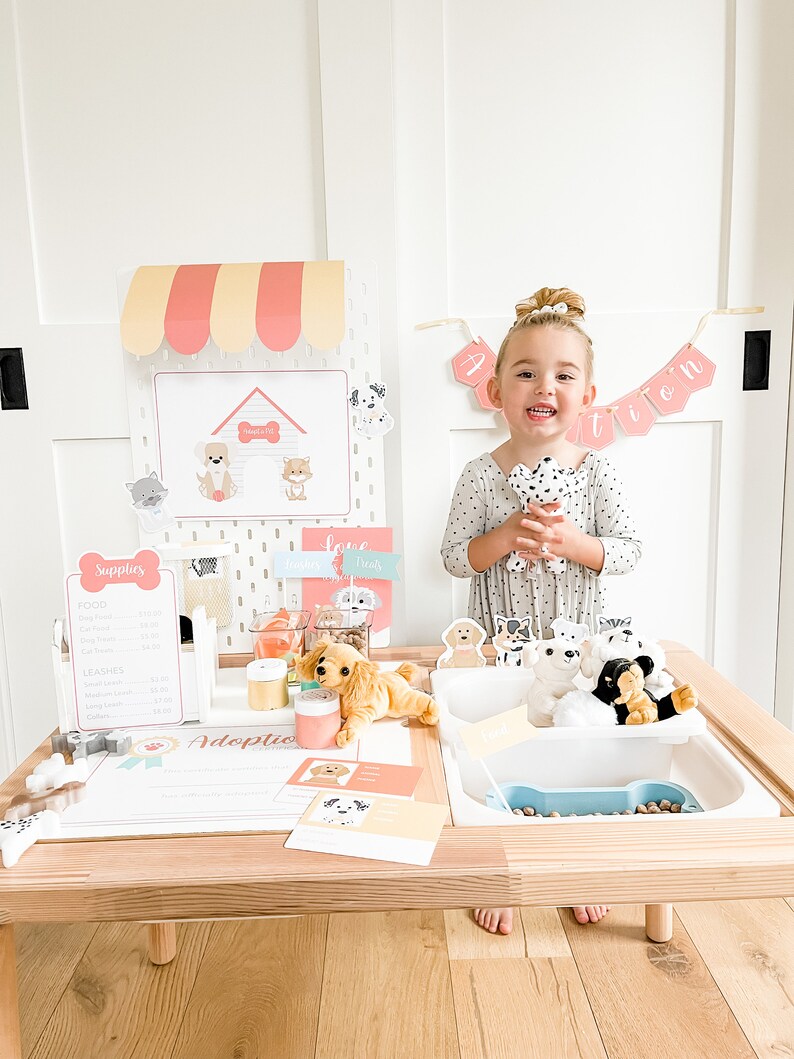 May include: A child plays with a pretend pet adoption booth. The booth has a sign that says "Adoption" and a variety of stuffed dogs. The child is holding a stuffed dog and smiling.