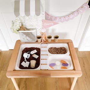 May include: A wooden sensory table with three white plastic inserts filled with various items. The table has a light wood frame and legs. A banner that says "Showers Flowers" hangs above the table. A small white metal basket and a decorative awning are also present.