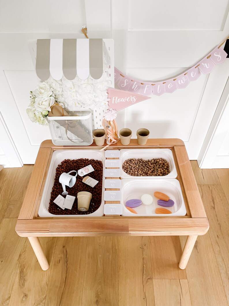 May include: A wooden sensory table with three white plastic inserts filled with various items. The table has a light wood frame and legs. A banner that says "Showers Flowers" hangs above the table. A small white metal basket and a decorative awning are also present.