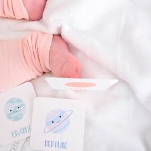 May include: A set of four flash cards featuring illustrations of planets with cartoon faces. The cards are white with blue and pink accents. The planets are Uranus, Neptune, Saturn, and Jupiter. The cards are on a white surface.