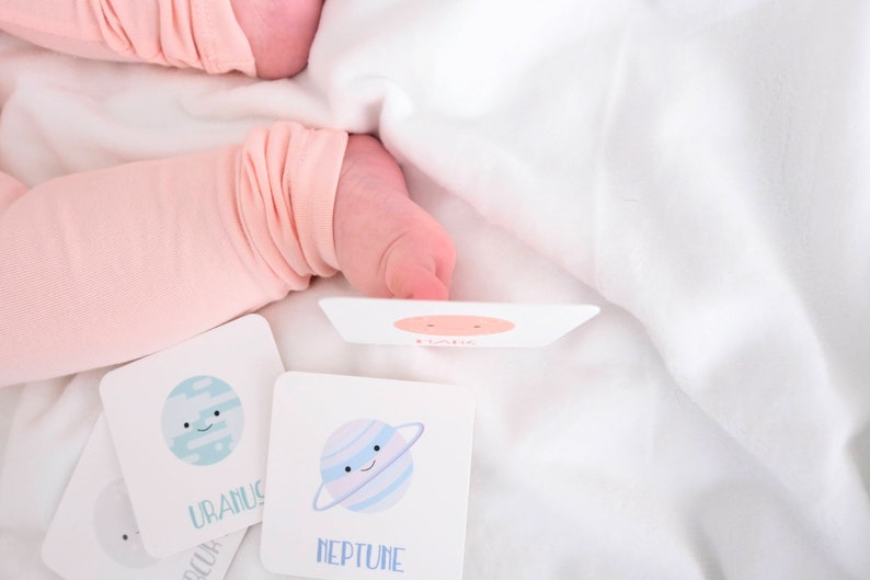 May include: A set of four flash cards featuring illustrations of planets with cartoon faces. The cards are white with blue and pink accents. The planets are Uranus, Neptune, Saturn, and Jupiter. The cards are on a white surface.