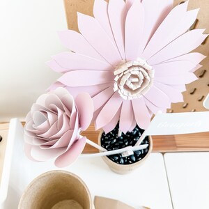 May include: A close-up of paper flowers in a small pot. The flowers are light pink and white, with a rose and a daisy. The pot is filled with black stones. A small sign reads "Bouquet".