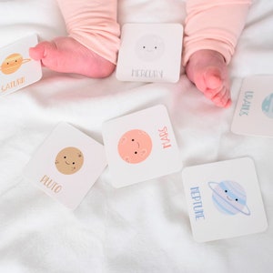 May include: A set of colorful flash cards featuring planets in our solar system. Each card has a cartoon illustration of a planet with a smiling face and the planet's name printed in white text. The cards are scattered on a white surface.