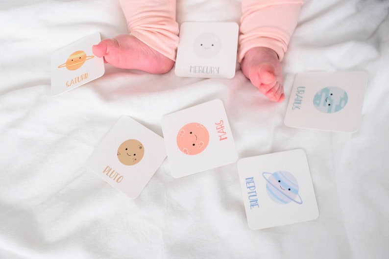 May include: A set of colorful flash cards featuring planets in our solar system. Each card has a cartoon illustration of a planet with a smiling face and the planet's name printed in white text. The cards are scattered on a white surface.