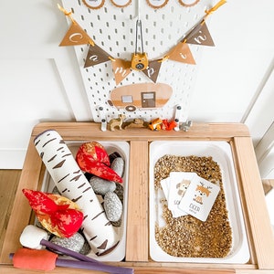 May include: A wooden play table with two white trays. One tray contains a faux campfire with red and orange flames, a birch log, and gray rocks. The other tray contains a brown sand-like material with two cards that say "Deer" and "Receive".