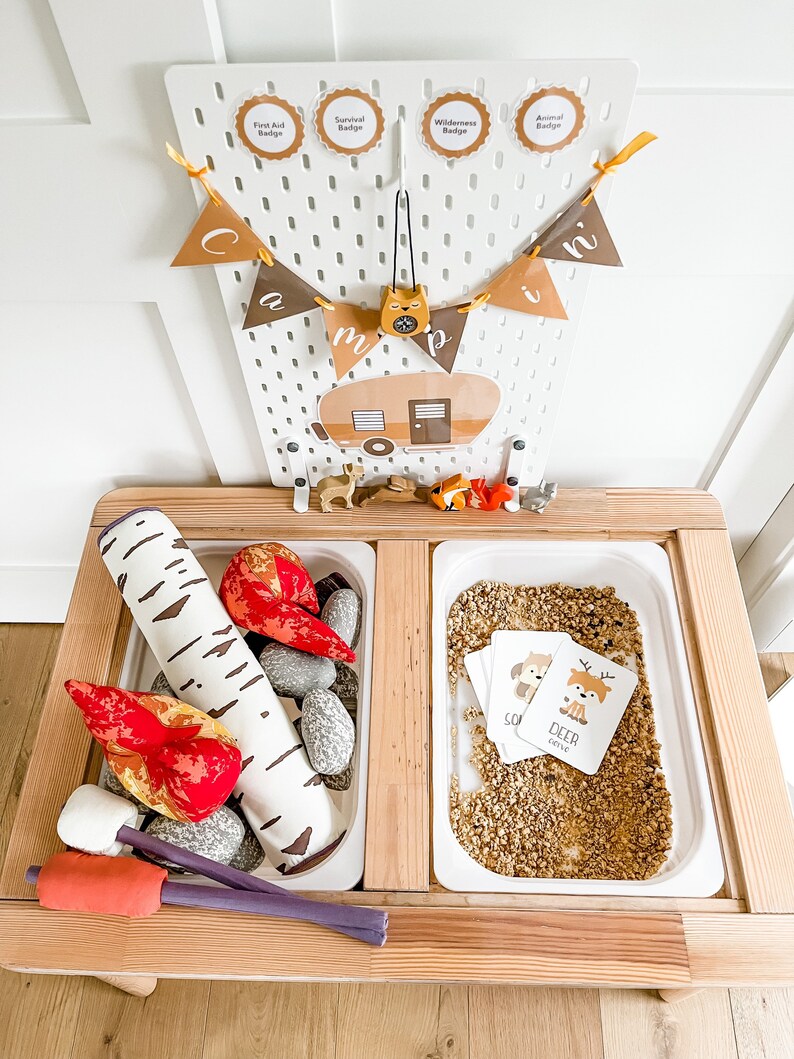 May include: A wooden play table with two white trays. One tray contains a faux campfire with red and orange flames, a birch log, and gray rocks. The other tray contains a brown sand-like material with two cards that say "Deer" and "Receive".
