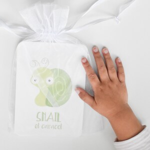 May include: A white organza bag containing a green and white illustrated card. The card depicts a cartoon snail with the words "Snail el caracol" printed on it.