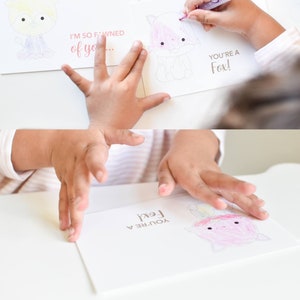 May include: Two blank Valentine's Day cards with a fox and a deer illustration. The cards have the text "You're a Fox!" and "I'm so fawned of you..." printed on them. A child is coloring the fox with a pink marker.