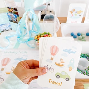 May include: A set of colorful flashcards with illustrations of travel-related items, such as a hot air balloon, airplane, sailboat, bicycle, and car. The cards have the word "Travel" printed on them in gold letters.