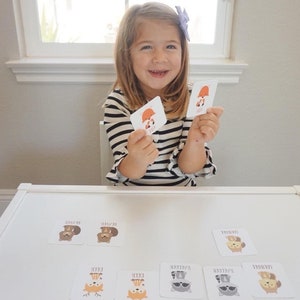 May include: A young girl is playing a matching game with cards featuring different woodland animals. The cards have illustrations of a fox, deer, raccoon, and bear. The cards also have the animal's name printed on them.