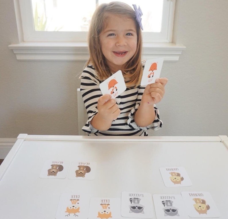 May include: A young girl is playing a matching game with cards featuring different woodland animals. The cards have illustrations of a fox, deer, raccoon, and bear. The cards also have the animal's name printed on them.