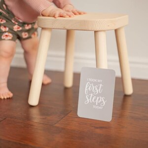 May include: A baby taking their first steps, holding onto a wooden stool. A gray card with the text "I took my first steps today" is on the floor.