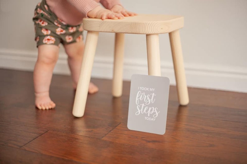 May include: A baby taking their first steps, holding onto a wooden stool. A gray card with the text "I took my first steps today" is on the floor.