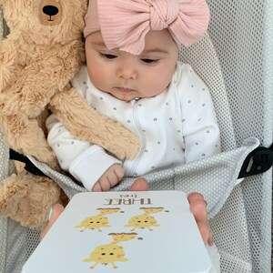 May include: A baby wearing a white onesie with a pink bow headband is holding a flashcard with the number three and the word "THREE" written on it. The flashcard features a cartoon illustration of three giraffes.