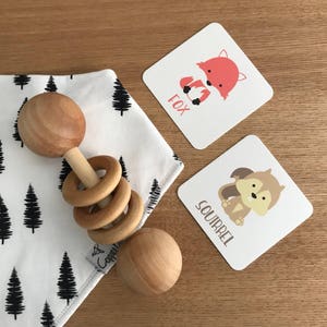 May include: A wooden baby rattle with a ring design and two round wooden balls. The rattle is on a white fabric with a black tree pattern. Two cards with animal illustrations are in the background. The cards say "Fox" and "Squirrel".