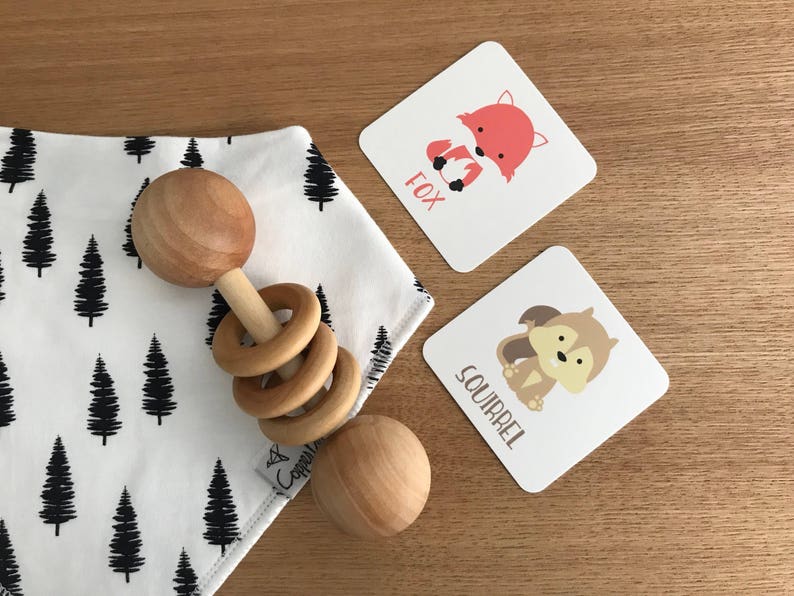 May include: A wooden baby rattle with a ring design and two round wooden balls. The rattle is on a white fabric with a black tree pattern. Two cards with animal illustrations are in the background. The cards say "Fox" and "Squirrel".