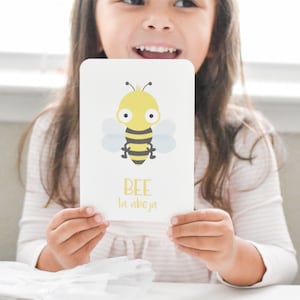 May include: A white card with a cartoon bee illustration and the words "BEE la abeja" in yellow text. A young girl is holding the card in front of her.