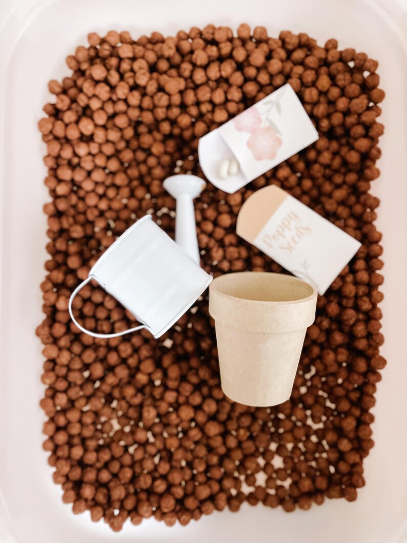 May include: A miniature gardening set featuring a small white watering can, a beige flower pot, and two seed packets. The set is surrounded by brown, spherical objects, possibly soil or mulch, within a white container.
