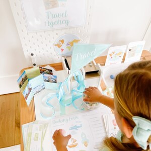 May include: A child plays with a pretend travel agency playset. The set includes a booking form, boarding passes, and a travel-themed flag. The child is wearing a light blue shirt and a white bow.