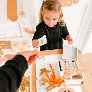 May include: A child plays with a toy pumpkin patch set, featuring a cash register, decorative pumpkins, and a menu board. The child is holding a small card, interacting with the play set. The scene is decorated with autumn-themed elements.