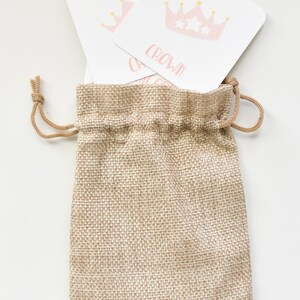 May include: A burlap drawstring bag with two white cards inside. The cards have pink crowns with gold stars on them. The text on the cards reads "CROWN".