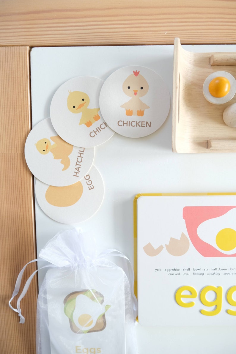 May include: A set of four colorful, illustrated learning cards featuring an egg, a chick hatching, a chick, and a chicken. The cards are on a white surface with a white organza bag containing a card with the word "Eggs" and an illustration of an egg on toast. The cards are part of a learning activity for children.
