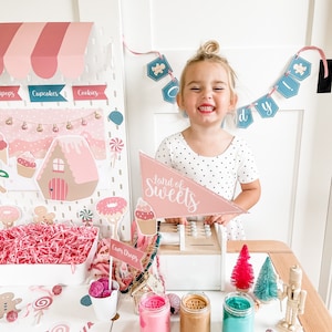 May include: A child plays pretend in a pink and white candy shop playset. The set includes a cash register, a sign that says "Land of Sweets", and various candy-themed props. The child is wearing a white polka dot dress and is smiling.