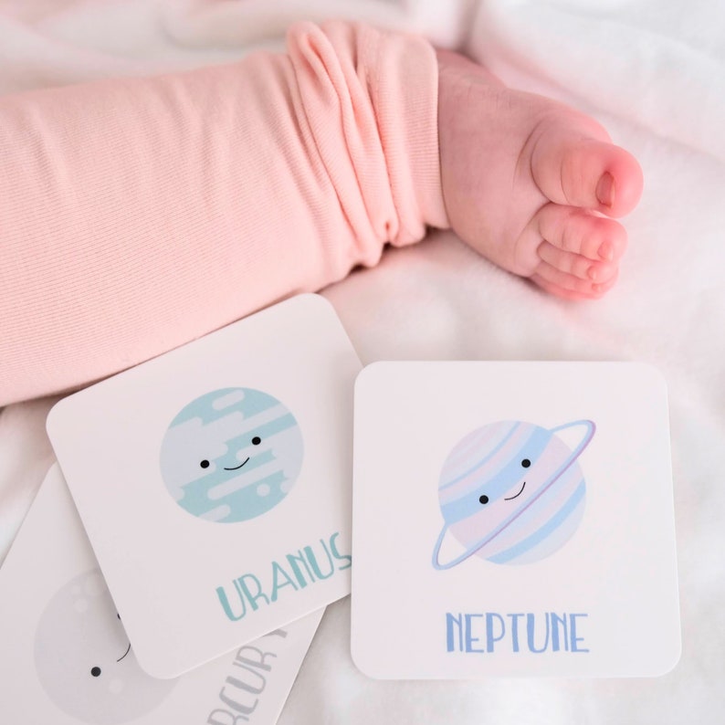May include: Four white cards with colorful illustrations of planets and their names. The cards feature Uranus, Neptune, Mercury, and Saturn. The cards are on a white surface with a baby's foot in the background.