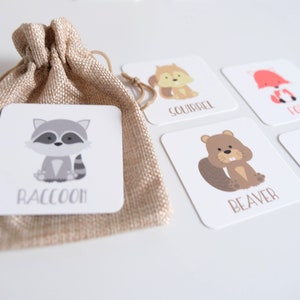 May include: Set of five illustrated flashcards featuring woodland animals. The cards are white with colorful illustrations of a raccoon, squirrel, fox, beaver, and deer. The name of each animal is printed below the illustration. The cards are in a small burlap bag.