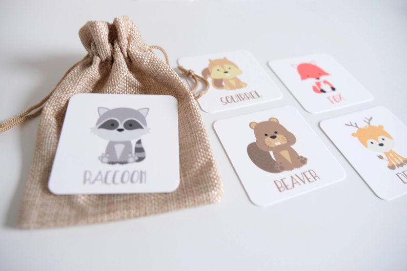 May include: Set of five illustrated flashcards featuring woodland animals. The cards are white with colorful illustrations of a raccoon, squirrel, fox, beaver, and deer. The name of each animal is printed below the illustration. The cards are in a small burlap bag.