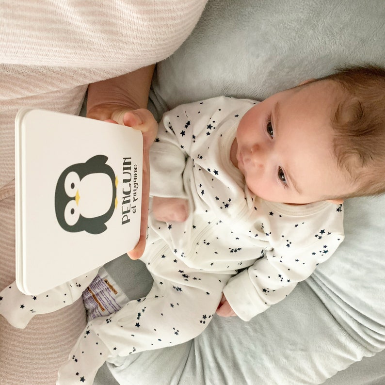 May include: A baby in a white onesie with black stars is looking at a white board book with a black and white penguin illustration. The book has the text "PENGUIN el ping&uuml;ino" on the cover.