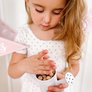 May include: A small white watering can filled with round, brown objects. The watering can has a spout with several holes. The person is holding the watering can with both hands. The person is wearing a white dress with black polka dots.