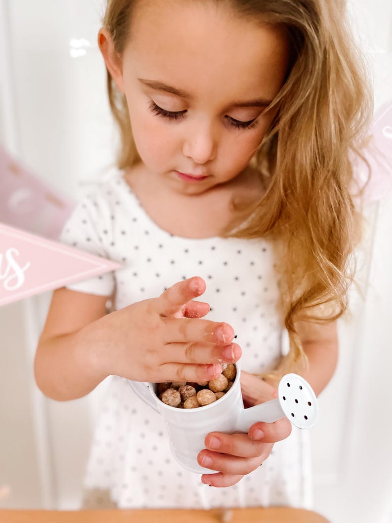 May include: A small white watering can filled with round, brown objects. The watering can has a spout with several holes. The person is holding the watering can with both hands. The person is wearing a white dress with black polka dots.