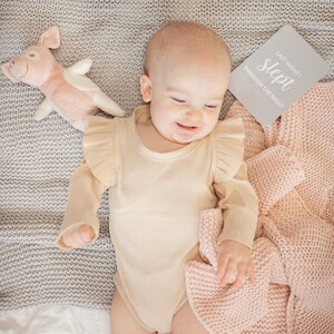 May include: A baby wearing a beige ribbed bodysuit with ruffled sleeves is lying on a gray knitted blanket. The baby is smiling and holding a pink knitted blanket. A pink pig stuffed animal is on the left side of the image. A white card with the text "Last night I slept through the night" is on the right side of the image.