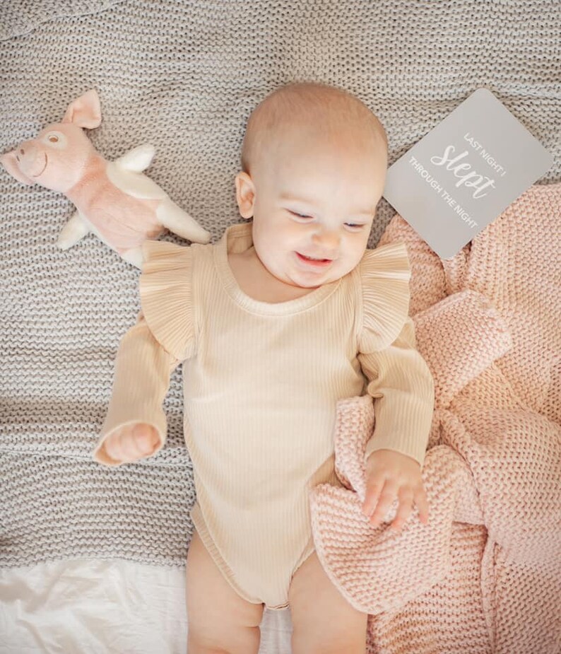 May include: A baby wearing a beige ribbed bodysuit with ruffled sleeves is lying on a gray knitted blanket. The baby is smiling and holding a pink knitted blanket. A pink pig stuffed animal is on the left side of the image. A white card with the text "Last night I slept through the night" is on the right side of the image.