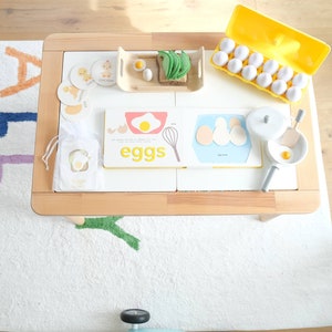 May include: A wooden table with a white rug underneath. On the table are various items related to eggs, including a book titled "eggs", a carton of plastic eggs, a small wooden tray with a toy egg, a toy pan with a toy egg, and a toy whisk. The rug has the word "DAILY" spelled out in colorful letters.