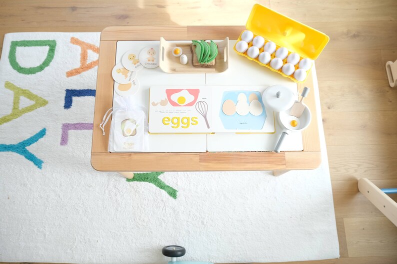 May include: A wooden table with a white rug underneath. On the table are various items related to eggs, including a book titled "eggs", a carton of plastic eggs, a small wooden tray with a toy egg, a toy pan with a toy egg, and a toy whisk. The rug has the word "DAILY" spelled out in colorful letters.