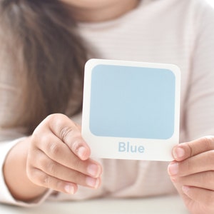 May include: A child holds up a blue color card with the word "Blue" printed on it. The card is part of a memory game for children ages 2 and up. The game is made by Goodnight Fox.
