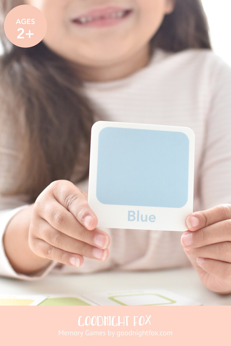 May include: A child holds up a blue color card with the word "Blue" printed on it. The card is part of a memory game for children ages 2 and up. The game is made by Goodnight Fox.