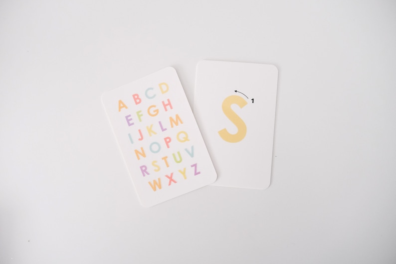 May include: Two white cards, one with the alphabet printed in colorful letters and the other with the letter "S" in yellow and a dotted line showing the direction to trace the letter.
