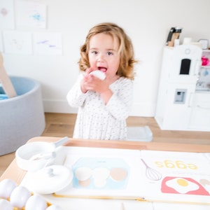 May include: A young child playing with toy eggs and a book about eggs. The book has a picture of a carton of eggs and a fried egg. The child is holding a white toy egg in their hand.