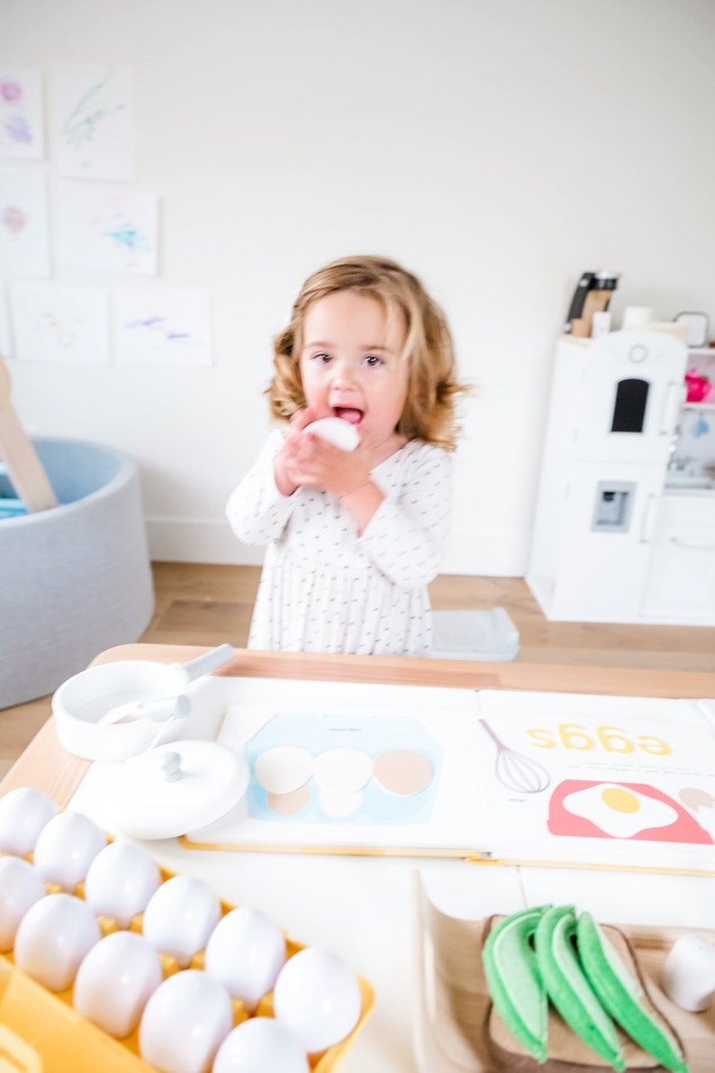 May include: A young child playing with toy eggs and a book about eggs. The book has a picture of a carton of eggs and a fried egg. The child is holding a white toy egg in their hand.